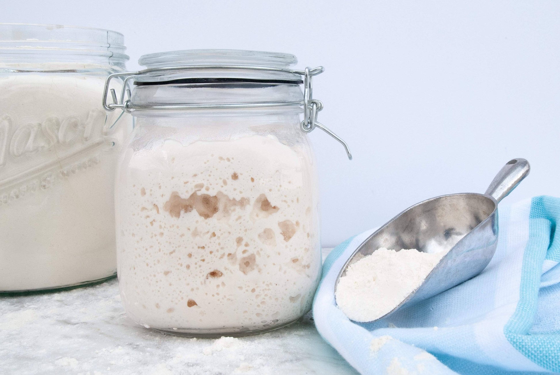 Jar of sourdough starter with a metal scoop on a light blue background