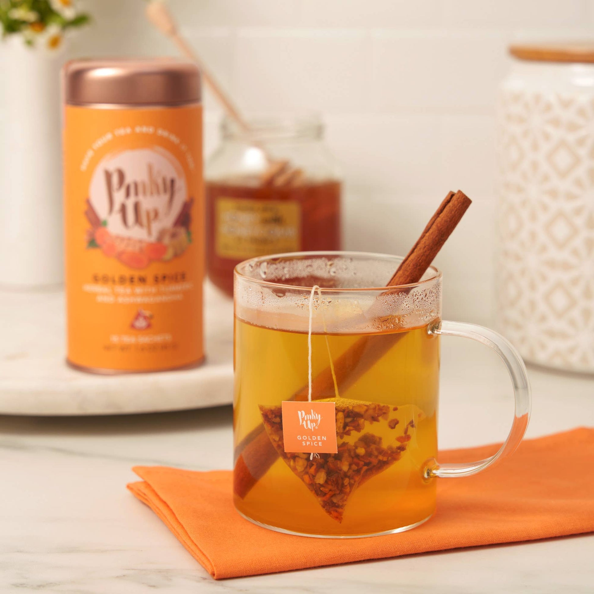 Glass mug of hot tea with cinnamon stick, 'Pinky Up' product in the background