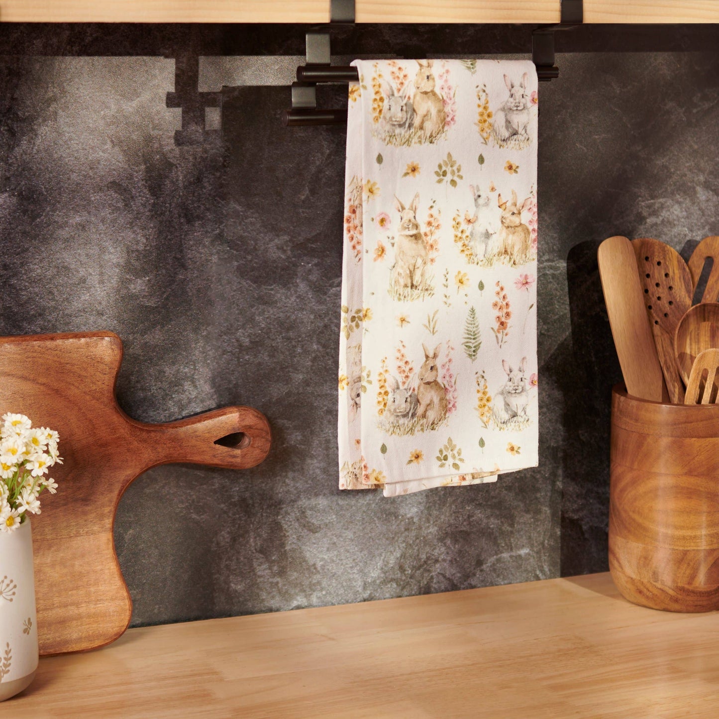 Kitchen counter with a floral towel, wooden cutting board, and utensils against a dark wall.