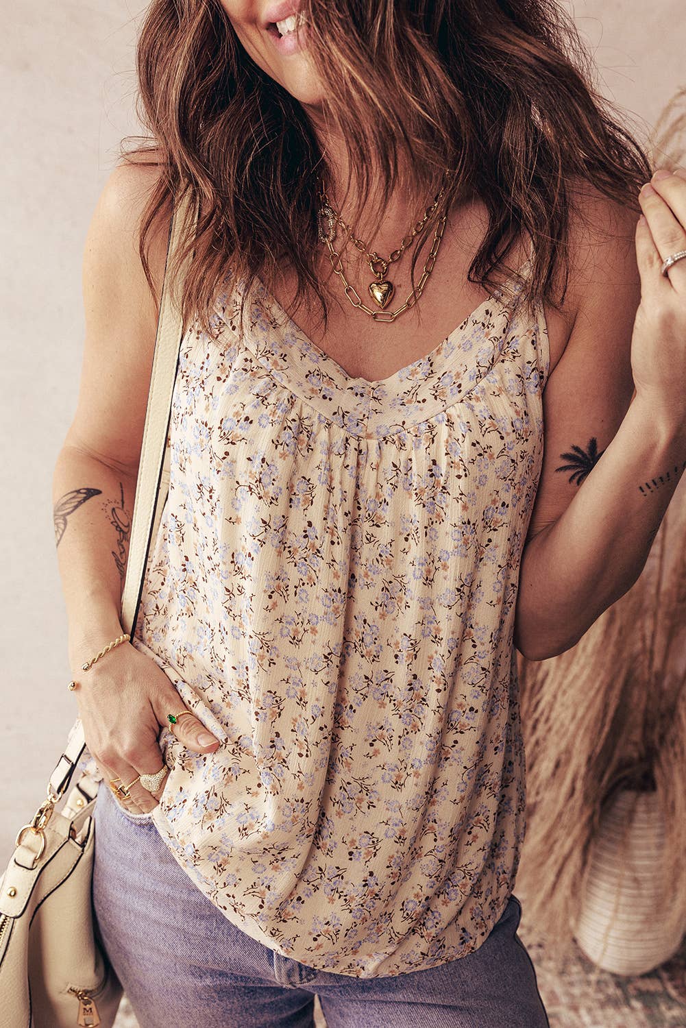 Woman wearing a floral sleeveless top with tattoos on her arms, standing against a neutral background.