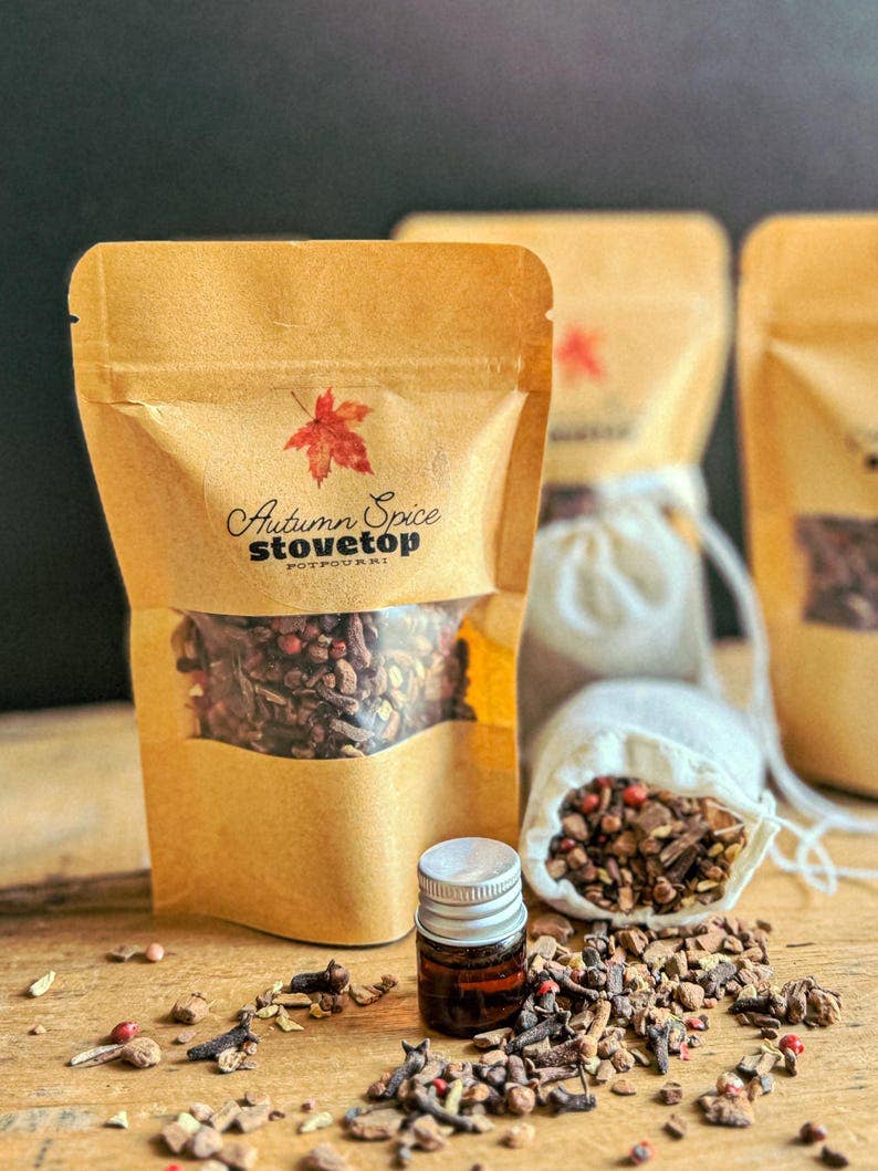 A stand-up kraft paper pouch labeled 'Autumn Spice stovetop' with a clear window showing loose spices inside, arranged on a wooden surface with additional spice bags, a small amber bottle, and scattered dried spices in front. The background is dark and softly blurred.
