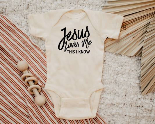 Baby onesie with 'Jesus loves me, this I know' text on a textured surface with a wooden toy.