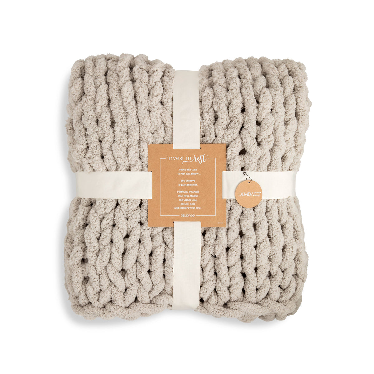 A chunky knit throw blanket in a warm oatmeal beige color, folded and wrapped with a cream ribbon and a tan paper tag. 