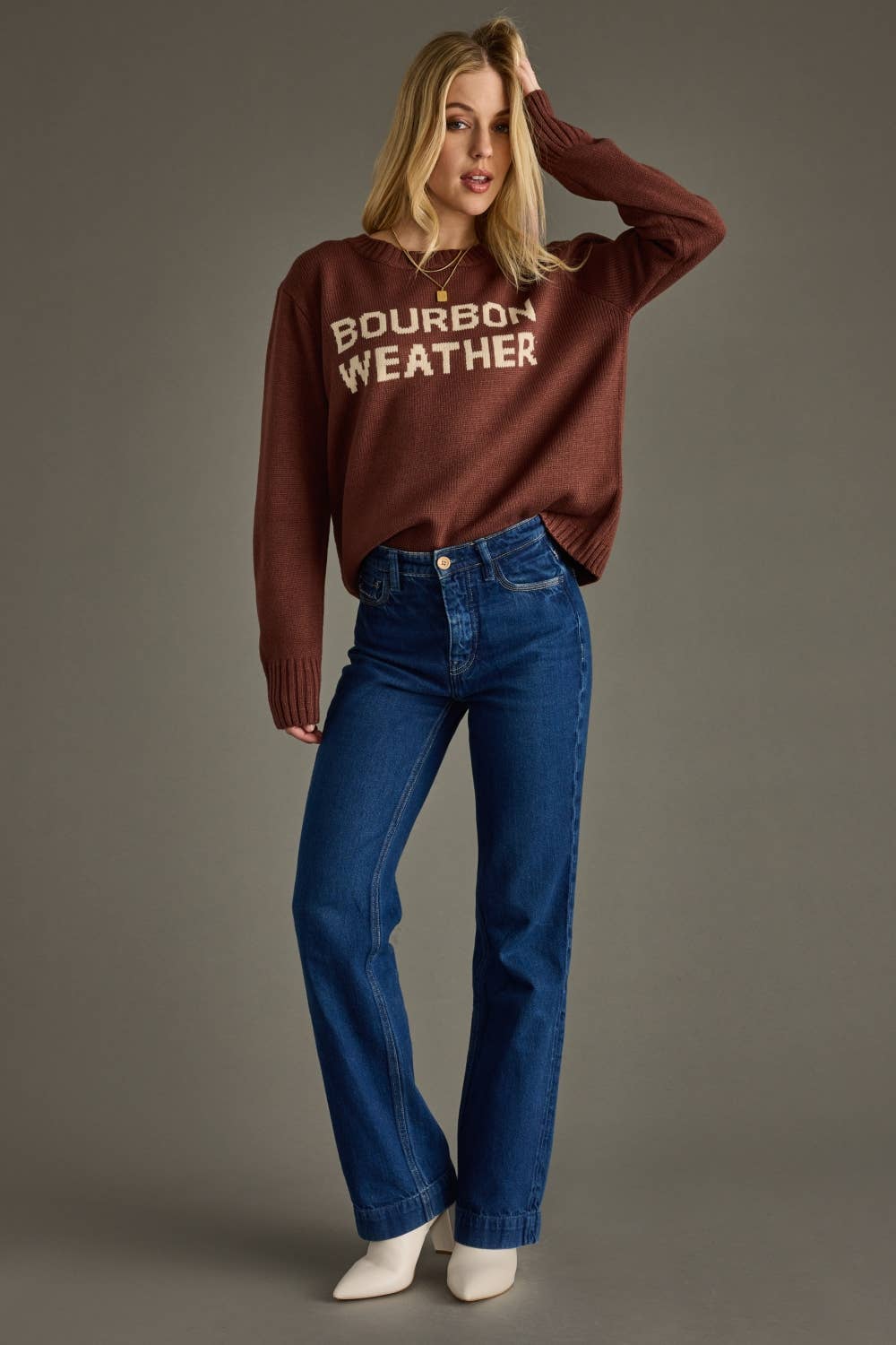 Woman wearing a brown sweater with 'Bourbon Weather' text and blue jeans on a gray background