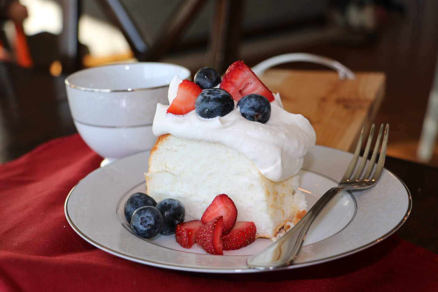 Slice of angel food cake with whipped cream and berries on a plate with a cup in the background.