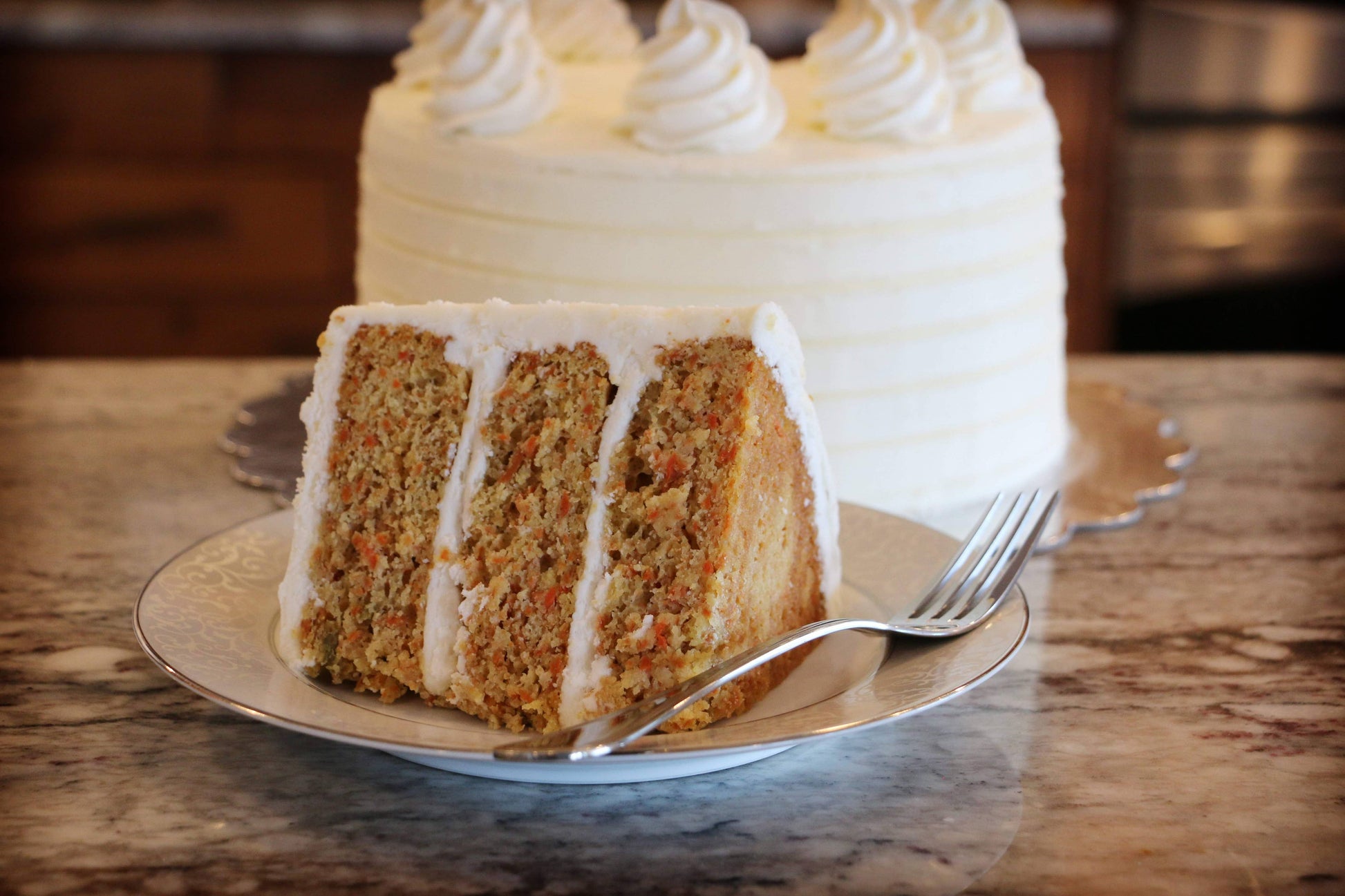 Carrot cake with white frosting on a plate with a fork, set against a kitchen background.