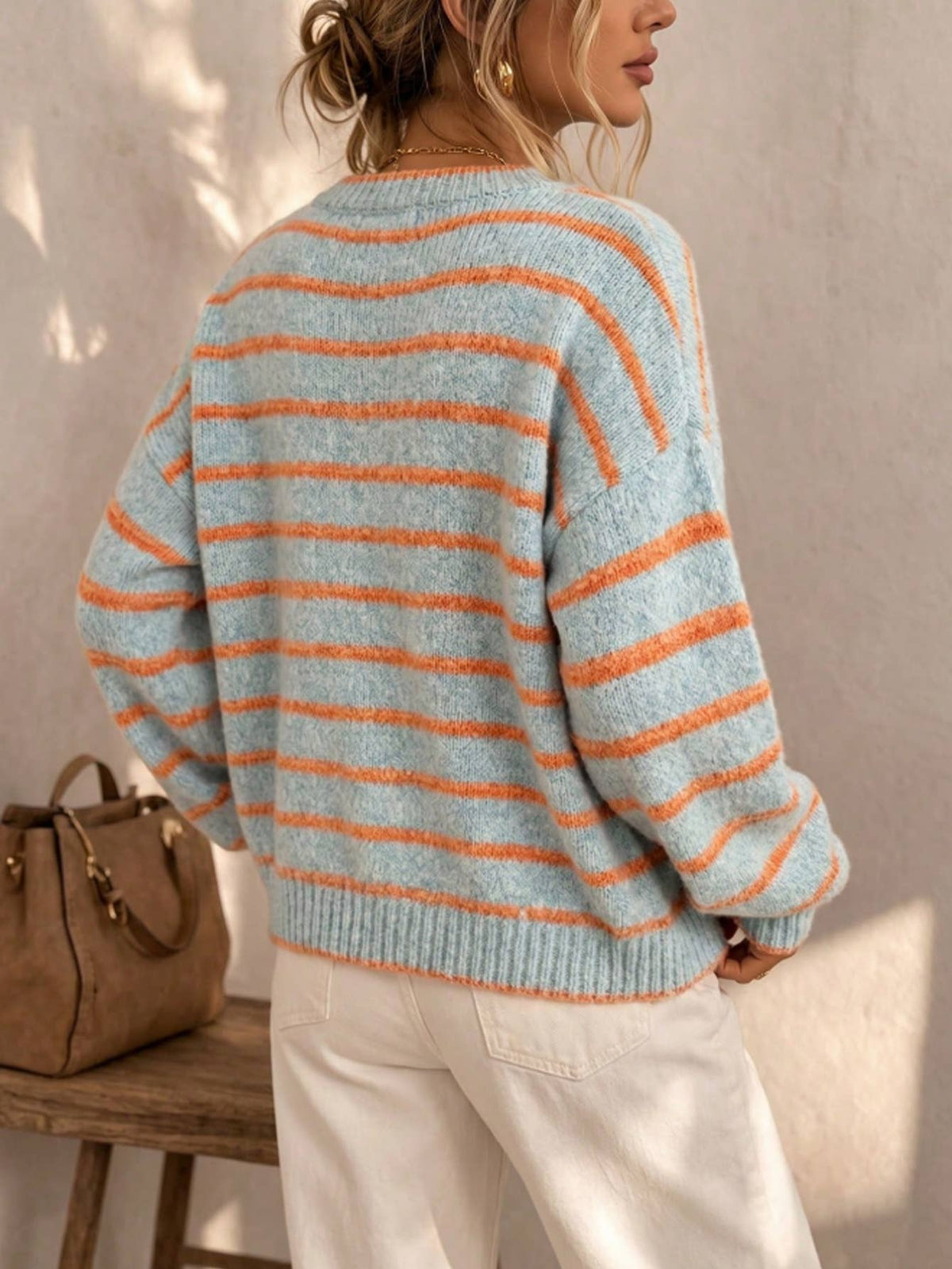 A woman wearing a light blue and orange striped knit sweater, shown from the back in a warm indoor setting with a beige wall and a brown handbag on a wooden bench.