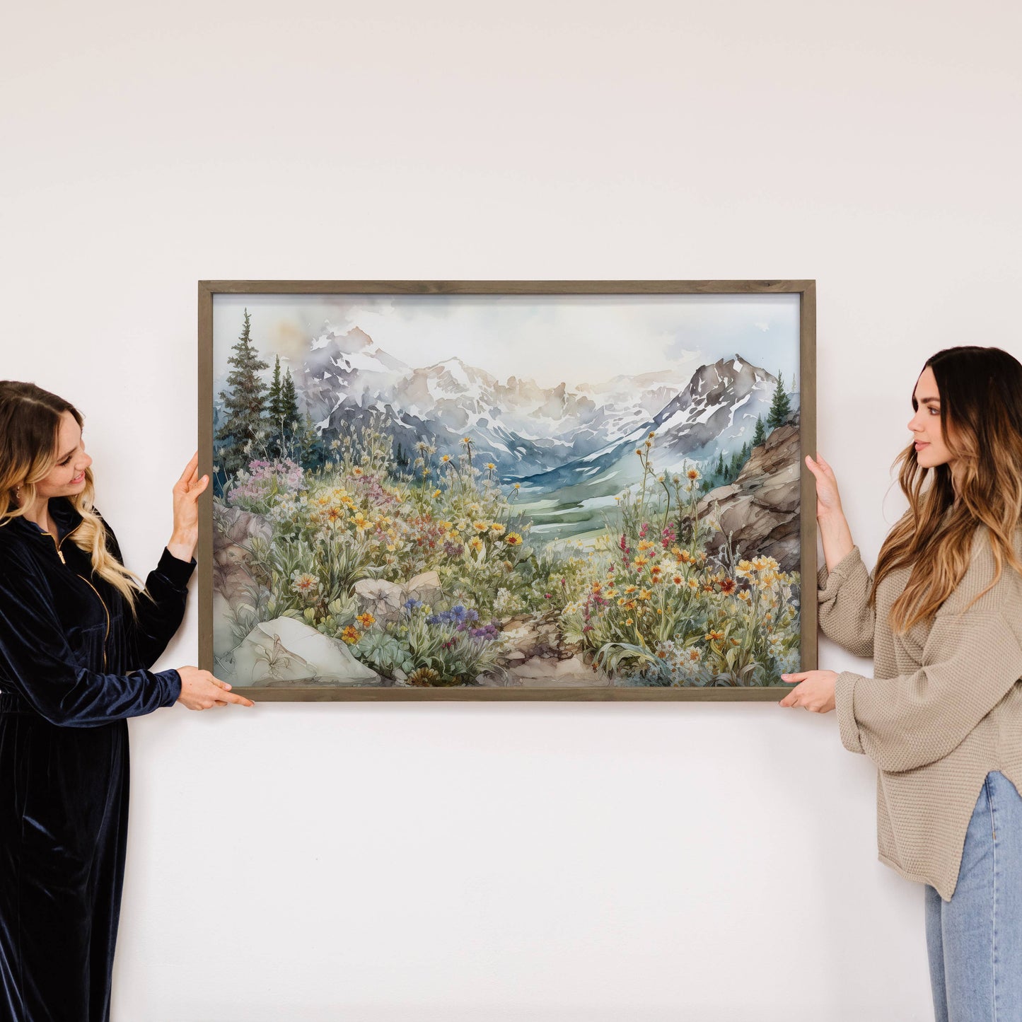 Two women holding a framed painting of a mountain landscape with flowers.