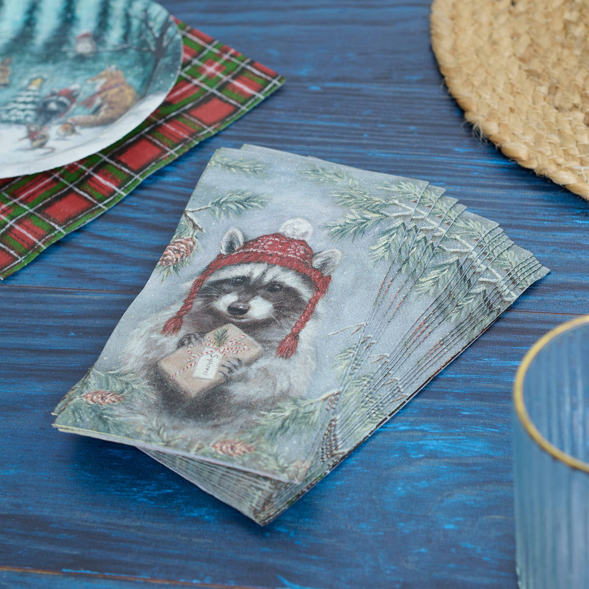 A stack of decorative paper napkins featuring a raccoon wearing a red knit hat and holding a gift, styled on a blue wooden tabletop with a plaid napkin, plate, woven mat, and glass partially visible.