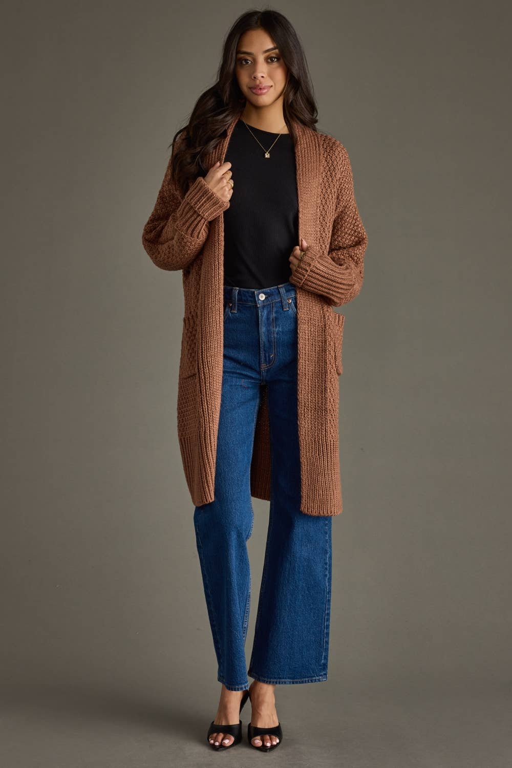 Woman wearing a long brown cardigan, black top, and blue jeans against a gray background
