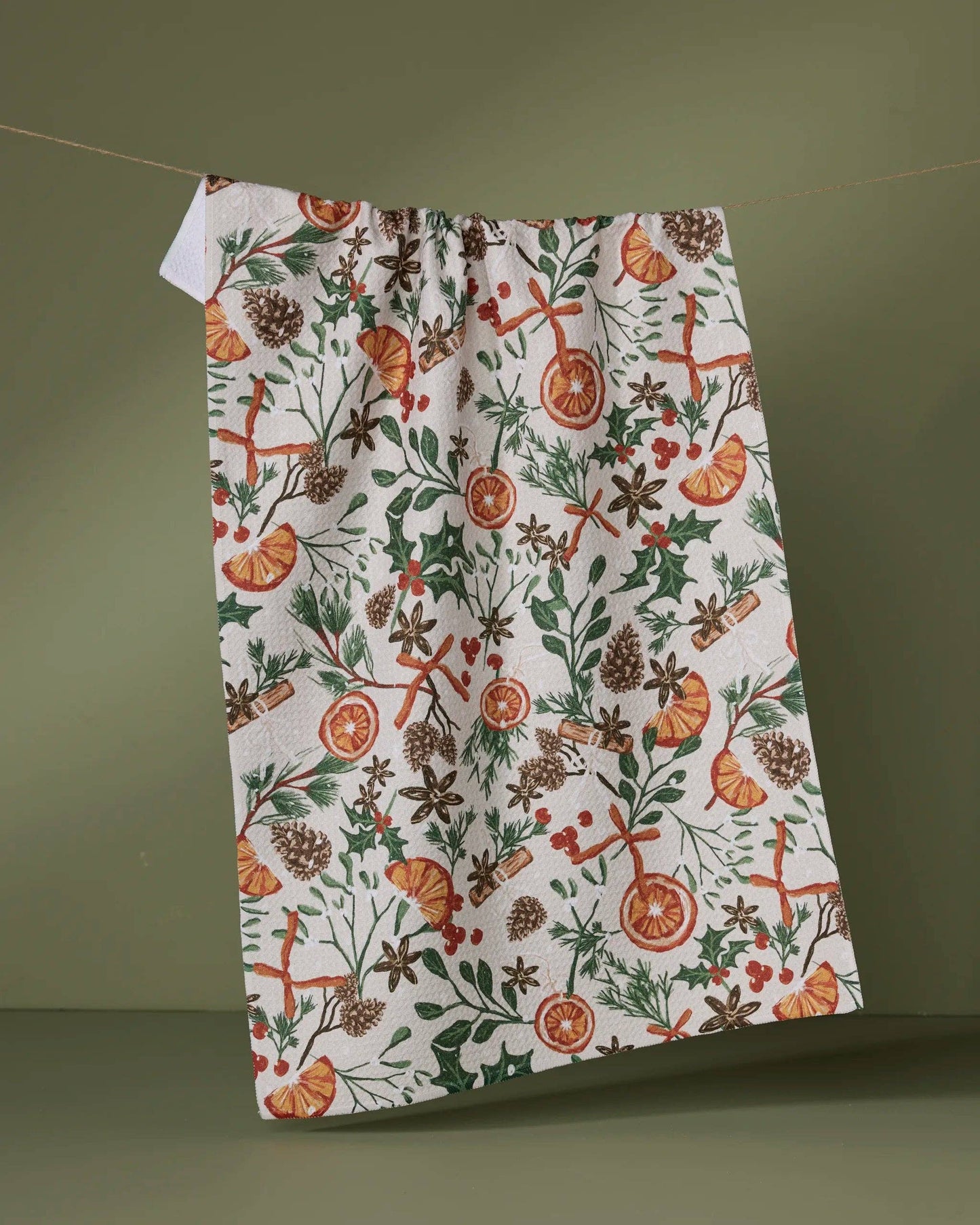 Geometry Tea Towel Canela  with a festive pattern of oranges, holly, and pine cones on a green background