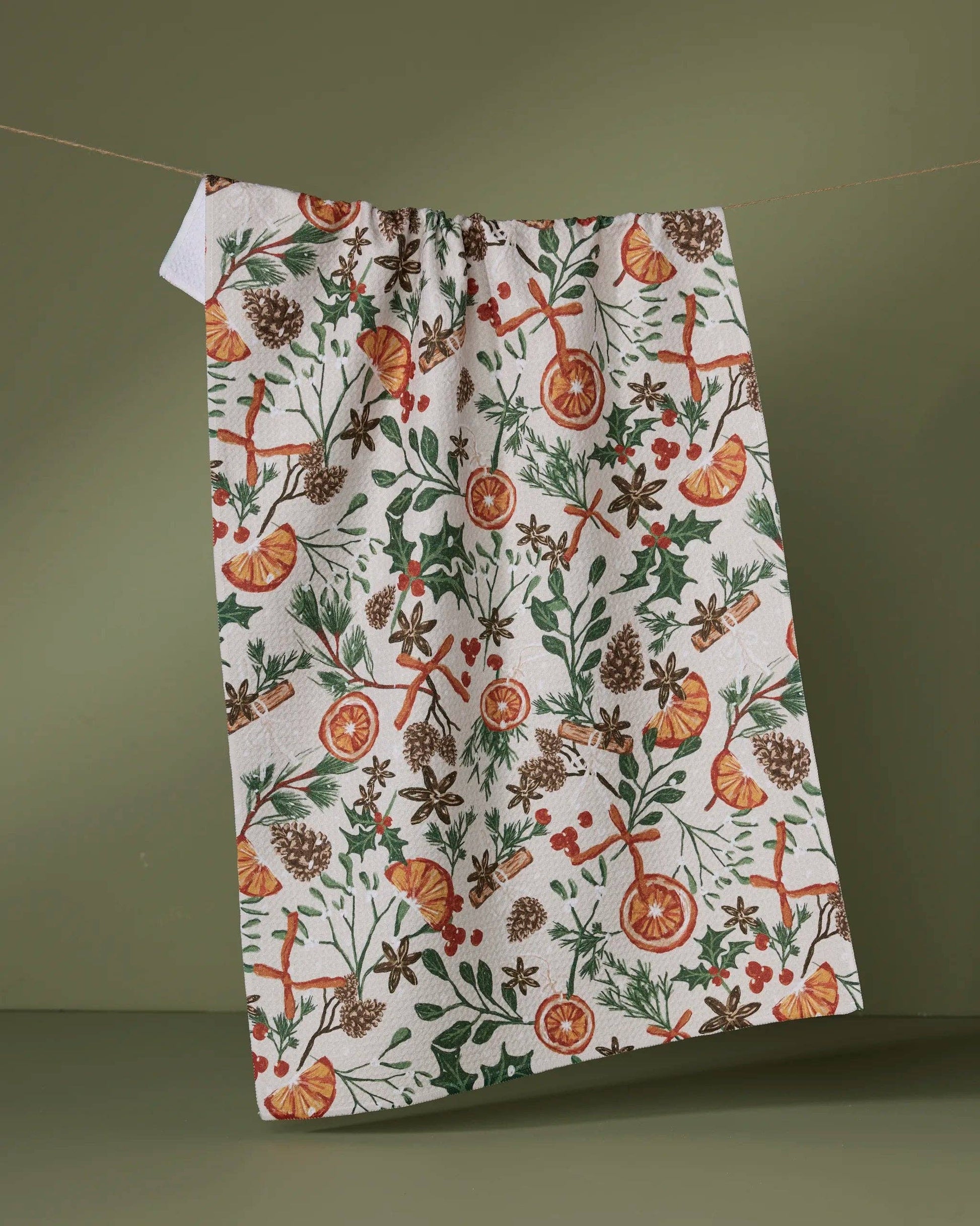 Geometry Tea Towel Canela  with a festive pattern of oranges, holly, and pine cones on a green background
