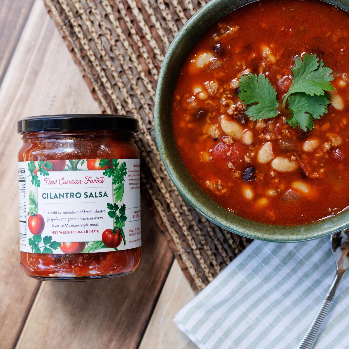 Jar of New Mexico Farms Cilantro Salsa next to a bowl of chili with beans and cilantro on a wooden surface.