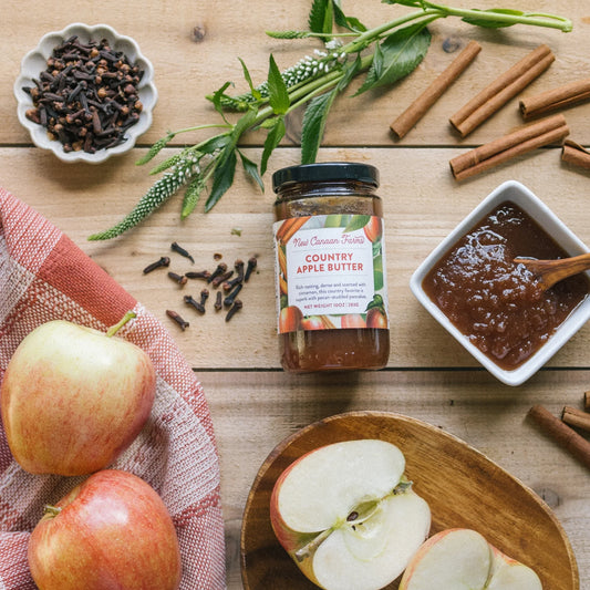 Jar of Country Apple Butter surrounded by apples, spices, and a bowl of apple butter on a wooden surface.