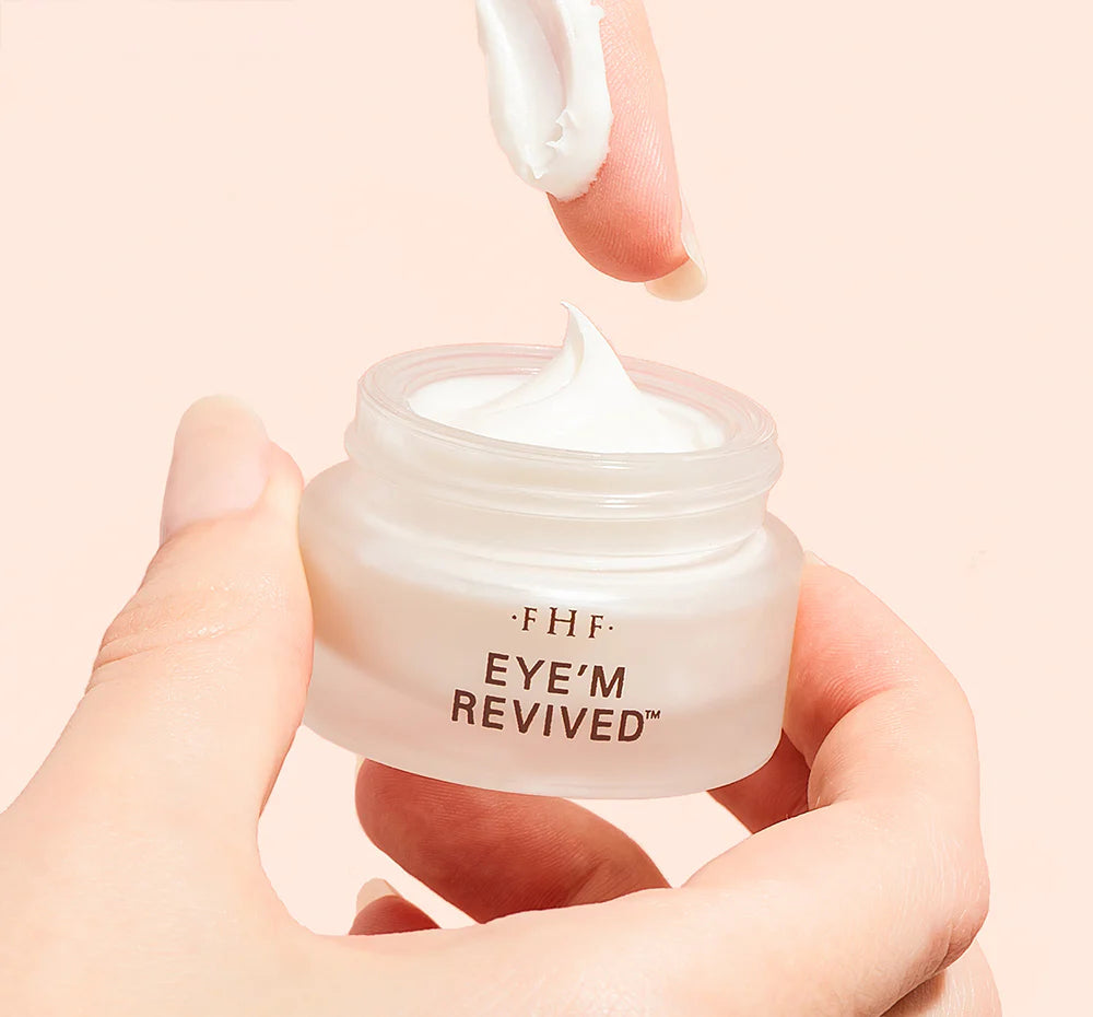 Hand holding a jar of 'EYE'M REVIVED' cream with a pink background