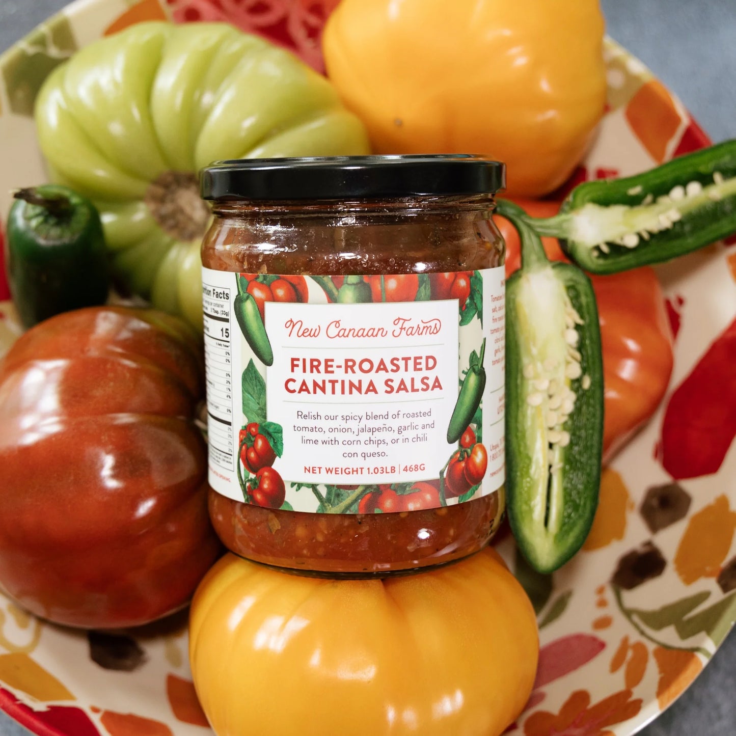 Jar of New Mexican Fire-Roasted Cantina Salsa surrounded by fresh tomatoes and jalapeños on a decorative plate.