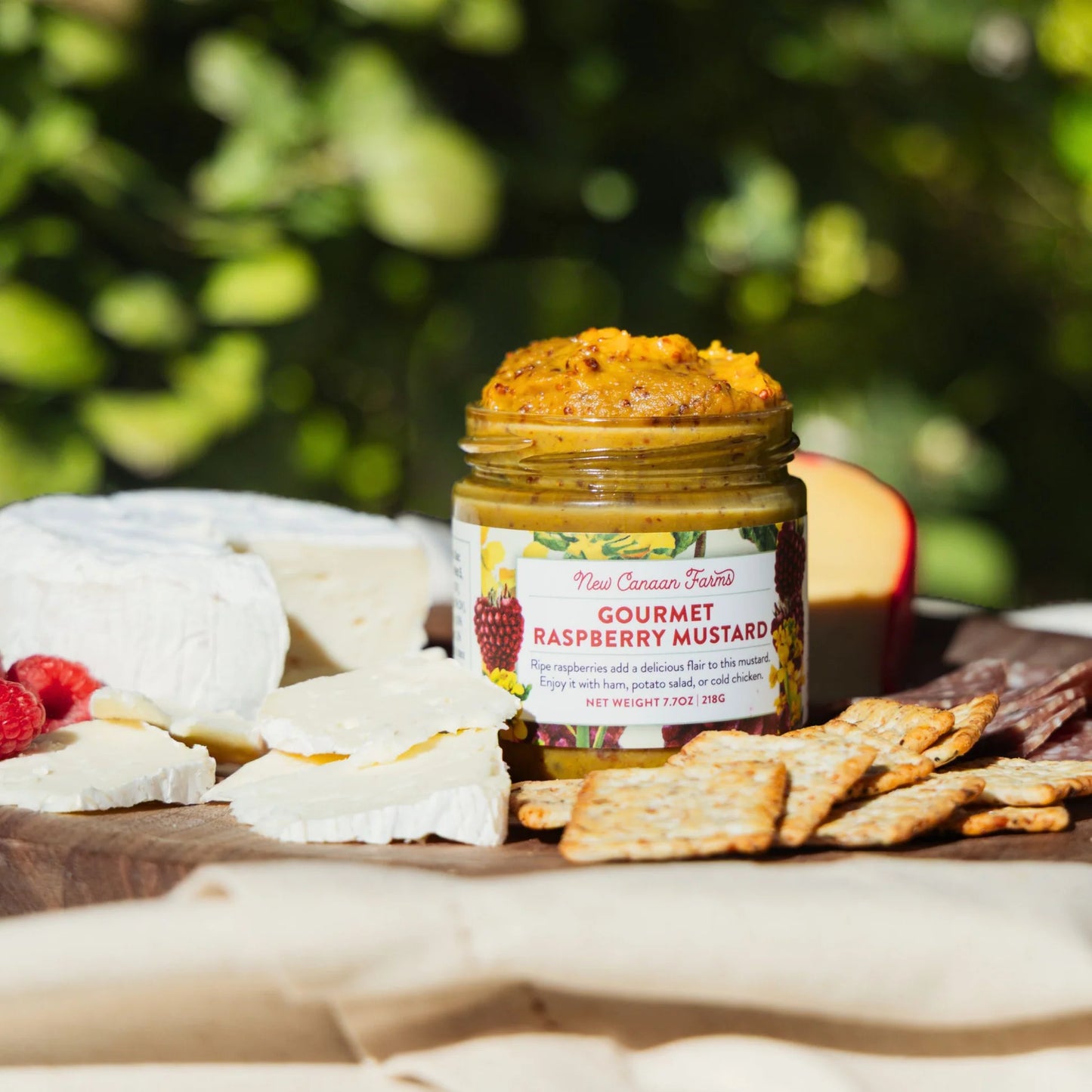 Jar of gourmet raspberry mustard on a wooden board with cheese and crackers outdoors.