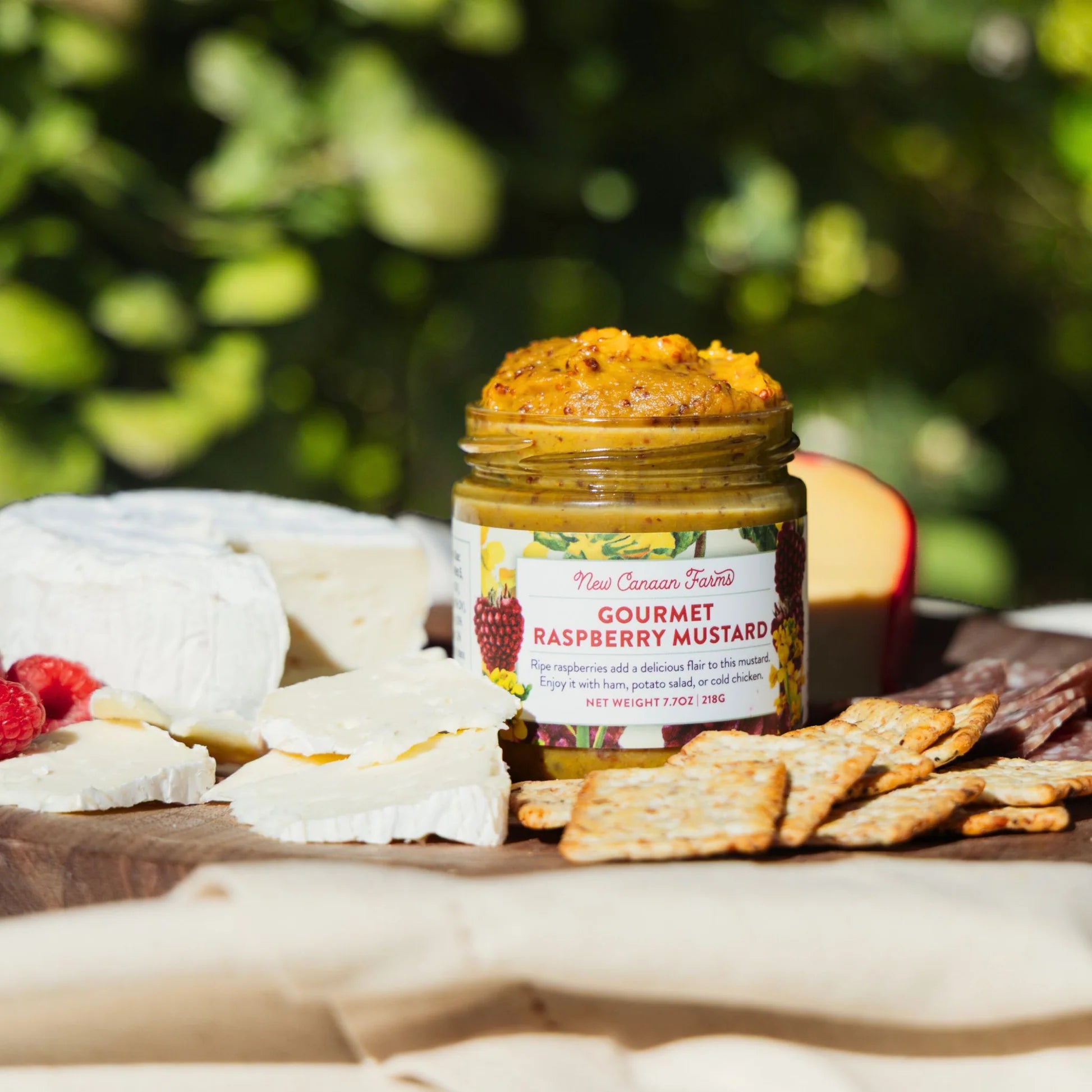 Jar of gourmet raspberry mustard on a wooden board with cheese and crackers outdoors.