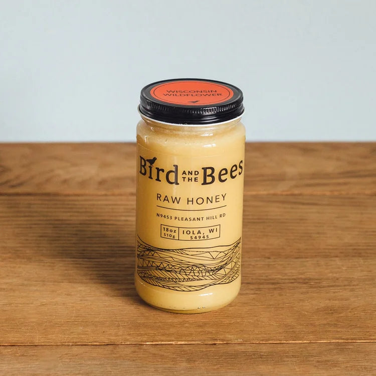Jar of 'Bird and the Bees' raw honey on a wooden surface with a light blue background