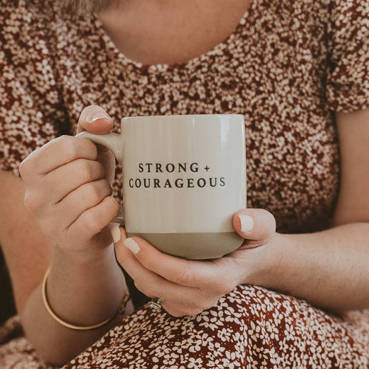 Person holding a mug with 'STRONG + COURAGEOUS' text