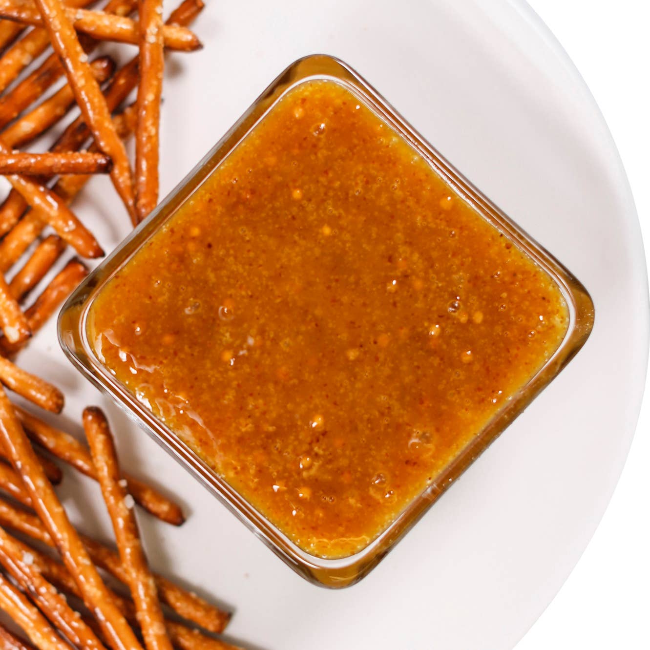 A square glass dipping sauce container filled with honey mustard sauce, placed on a white plate next to pretzel sticks.