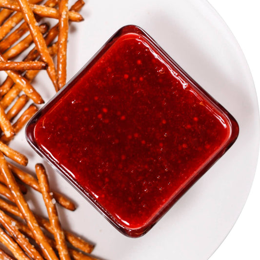 A square glass bowl filled with red raspberry honey mustard dip, placed on a white plate next to pretzel sticks.