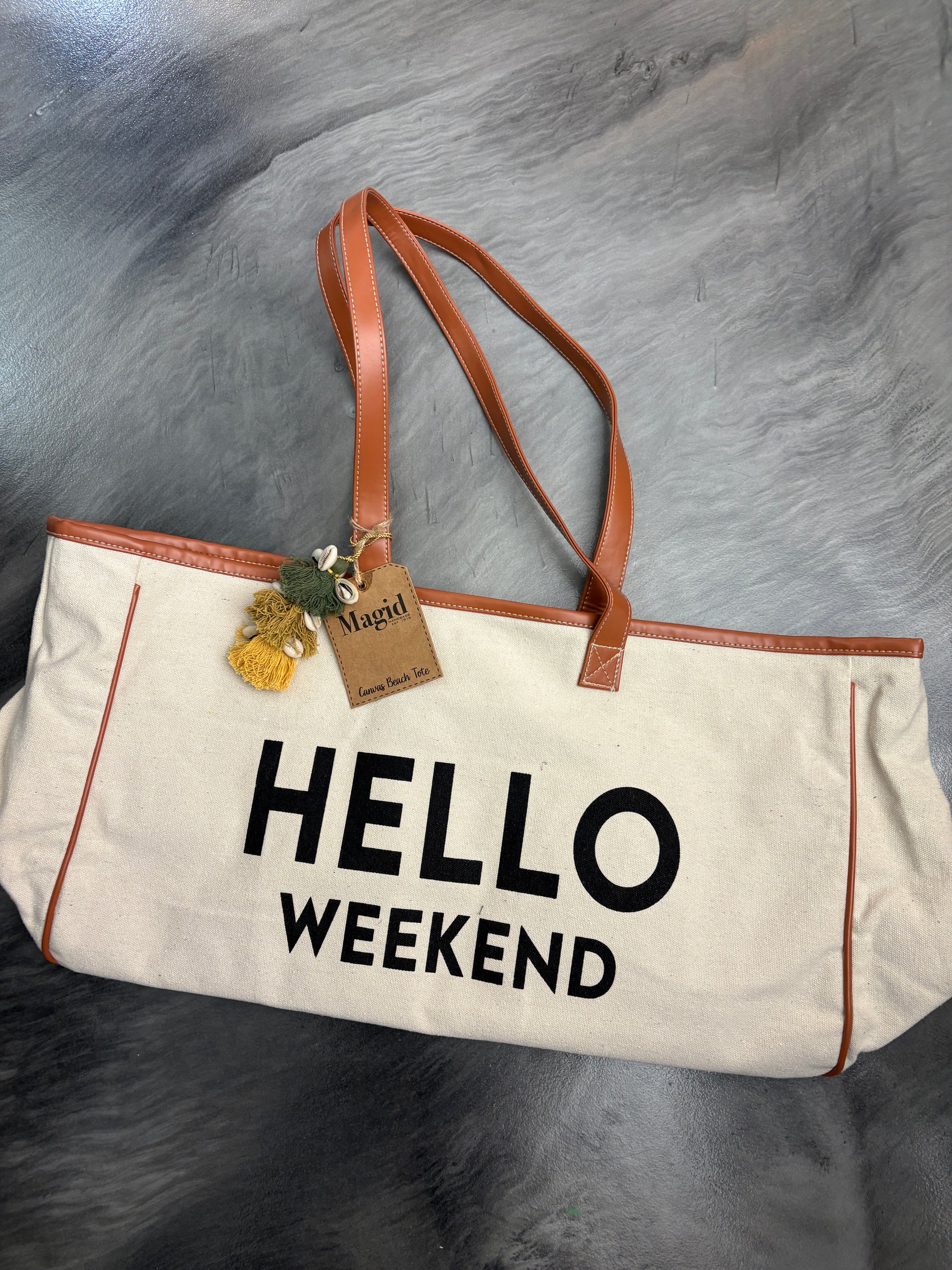 Beige tote bag with 'HELLO WEEKEND' text on a textured gray surface