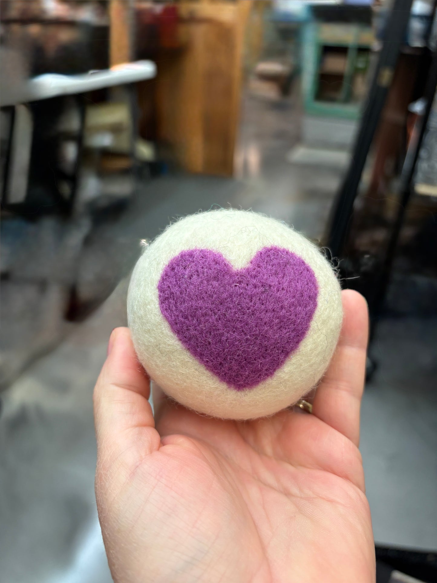 Hand holding a white wool dryer ball with a purple heart design against a blurred indoor background