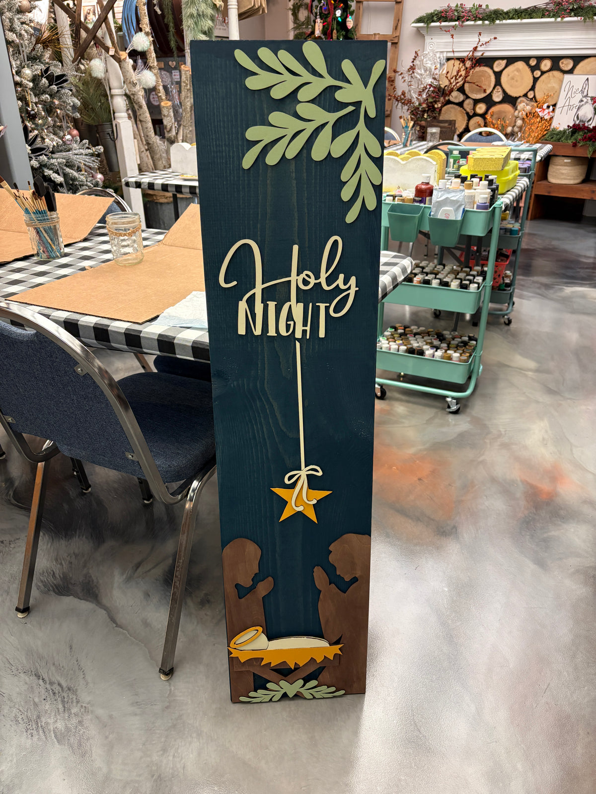 A tall rectangular wooden sign with a dark teal background, featuring a light green leafy branch at the top, the words 'Holy Night' in white script, a gold star, and brown hands holding a gold and white baby figure at the bottom. The sign is photographed standing on a gray concrete floor inside a craft store or workshop.
