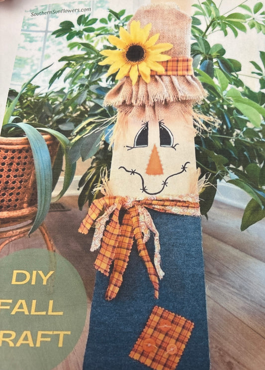 Scarecrow-themed DIY fall craft with a sunflower decoration against a plant background.