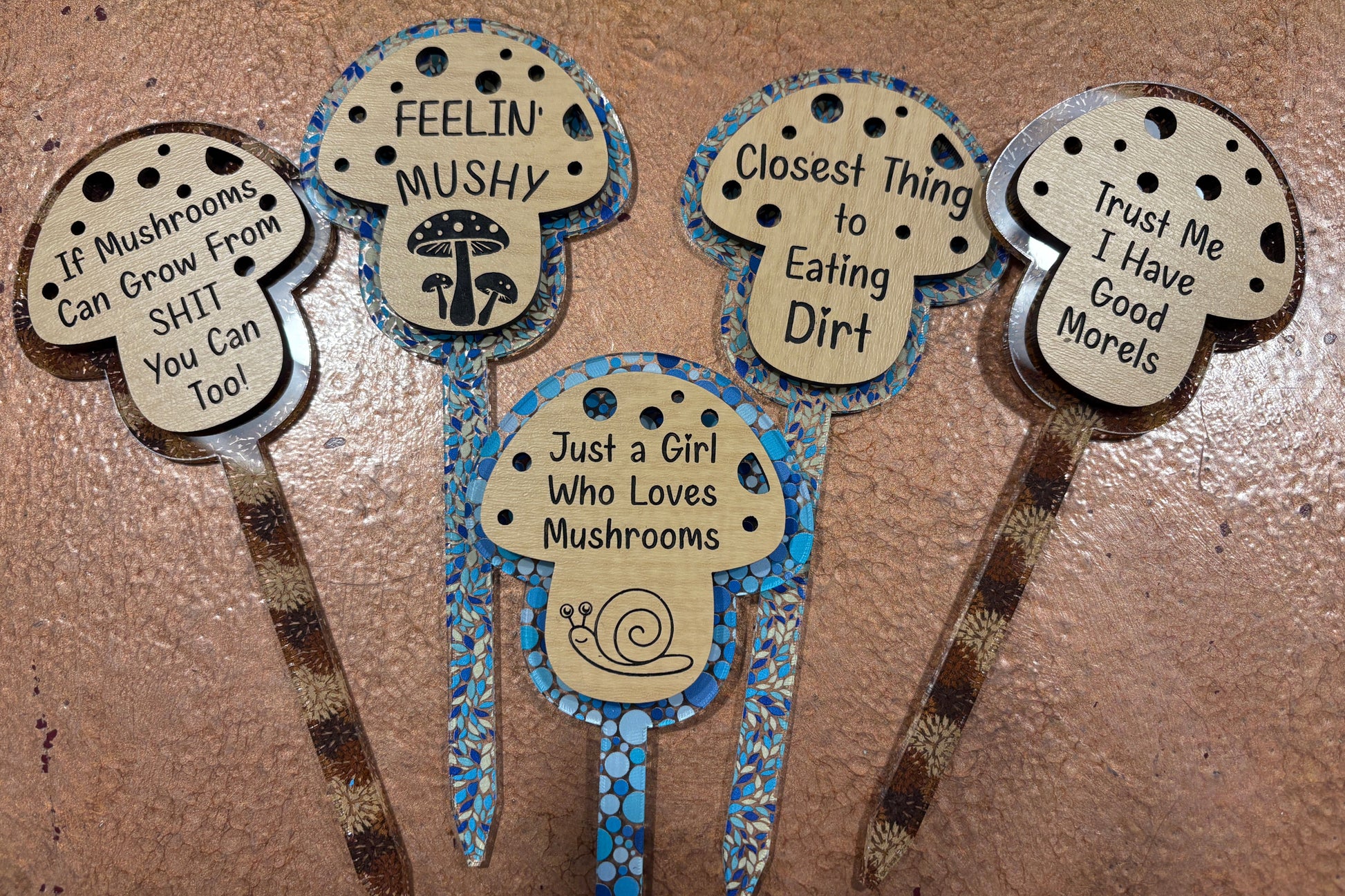 Five decorative mushroom-themed garden stakes with humorous sayings on a textured surface.
