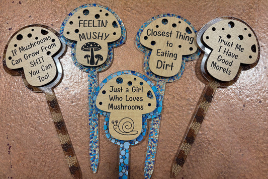 Five decorative mushroom-themed garden stakes with humorous sayings on a textured surface.