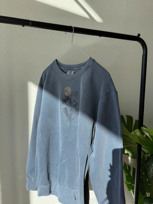Comfort Color Gray Crewneck Sweatshirt with Wolf & Moon embroidered on it hanging on a wire hanger and black rod on white background with green plant in the background