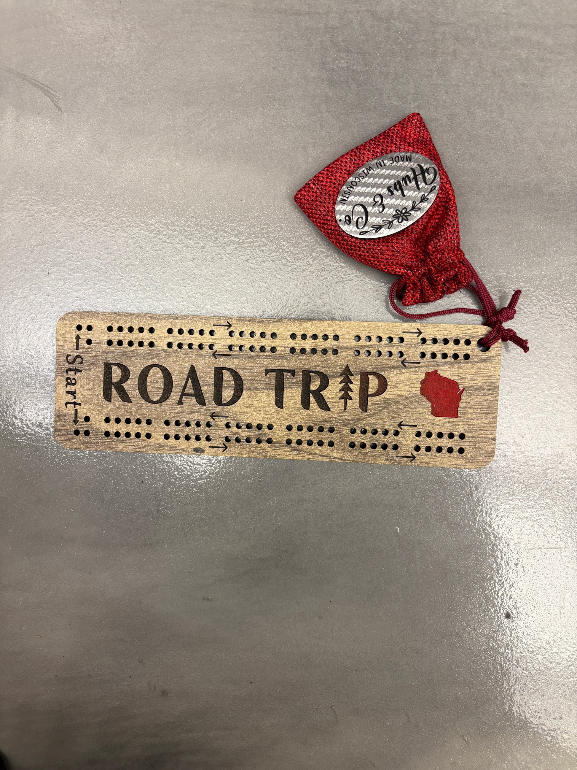 Wooden cribbage board with 'ROAD TRIP Start' text and red drawstring bag on a metallic surface