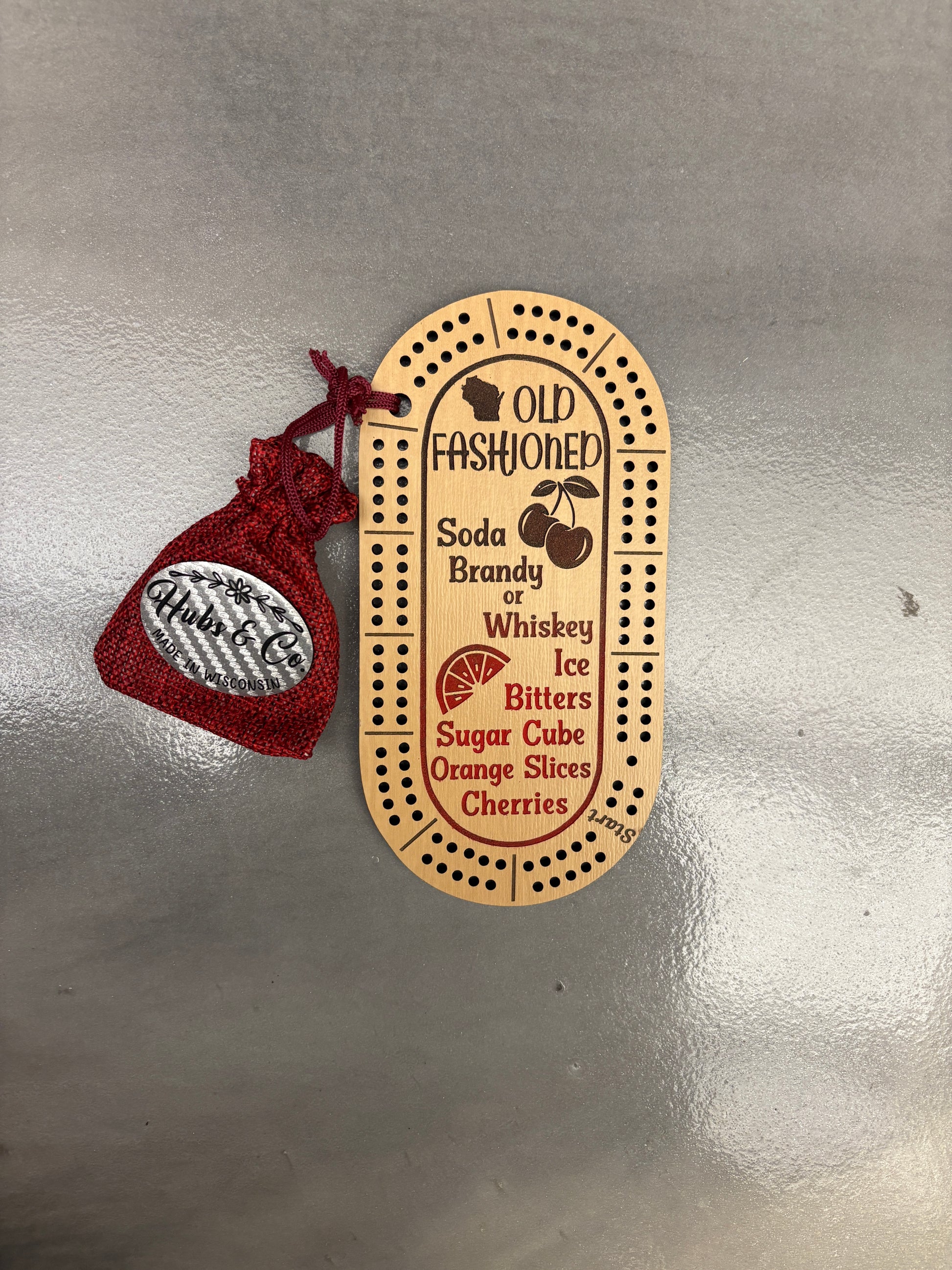 Wooden Old Fashioned cocktail recipe on mini cribbage board with a red drawstring on a metallic surface