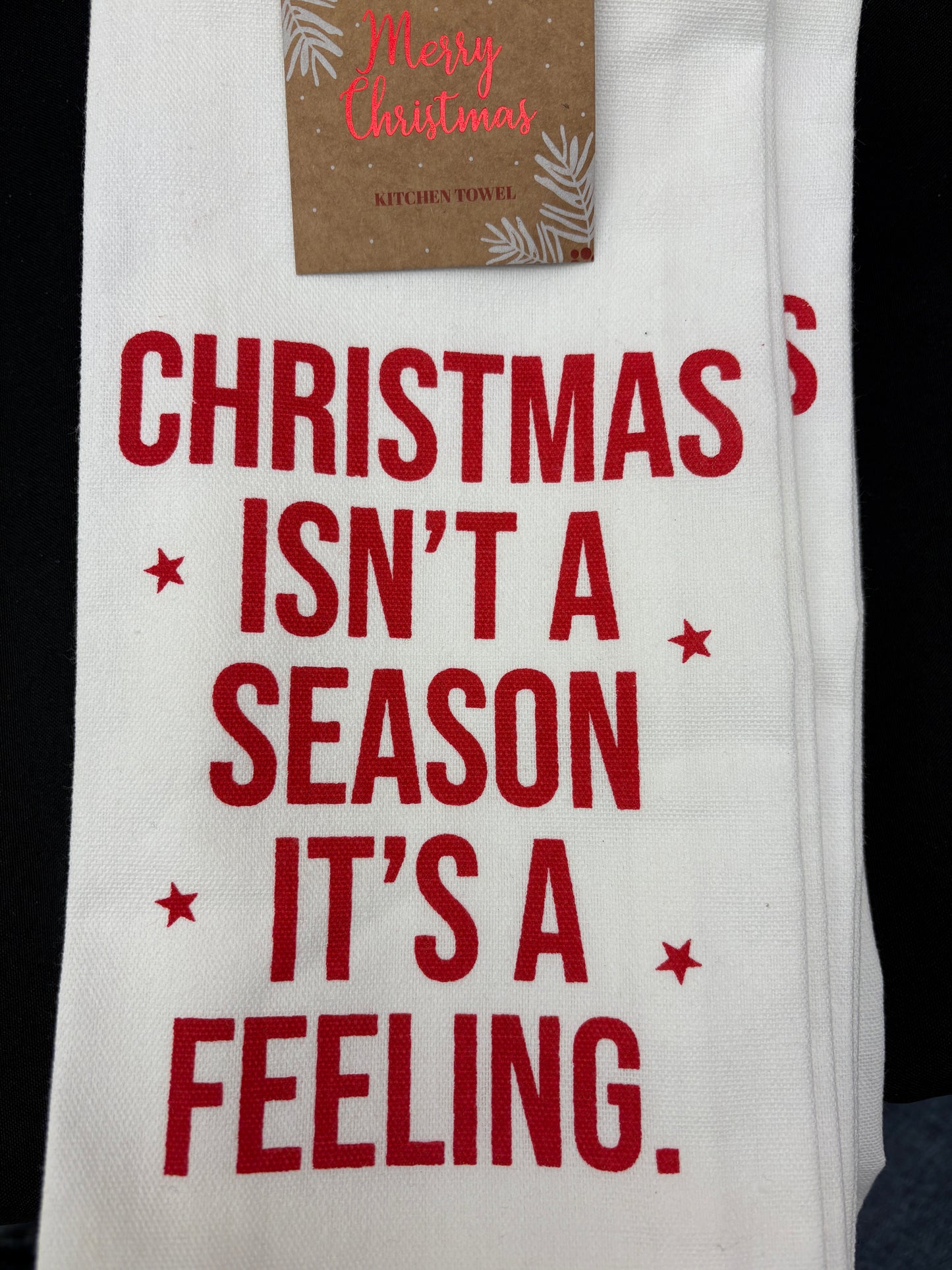 White towel with red text 'Christmas isn't a season, it's a feeling.' and a small brown tag on a black background.