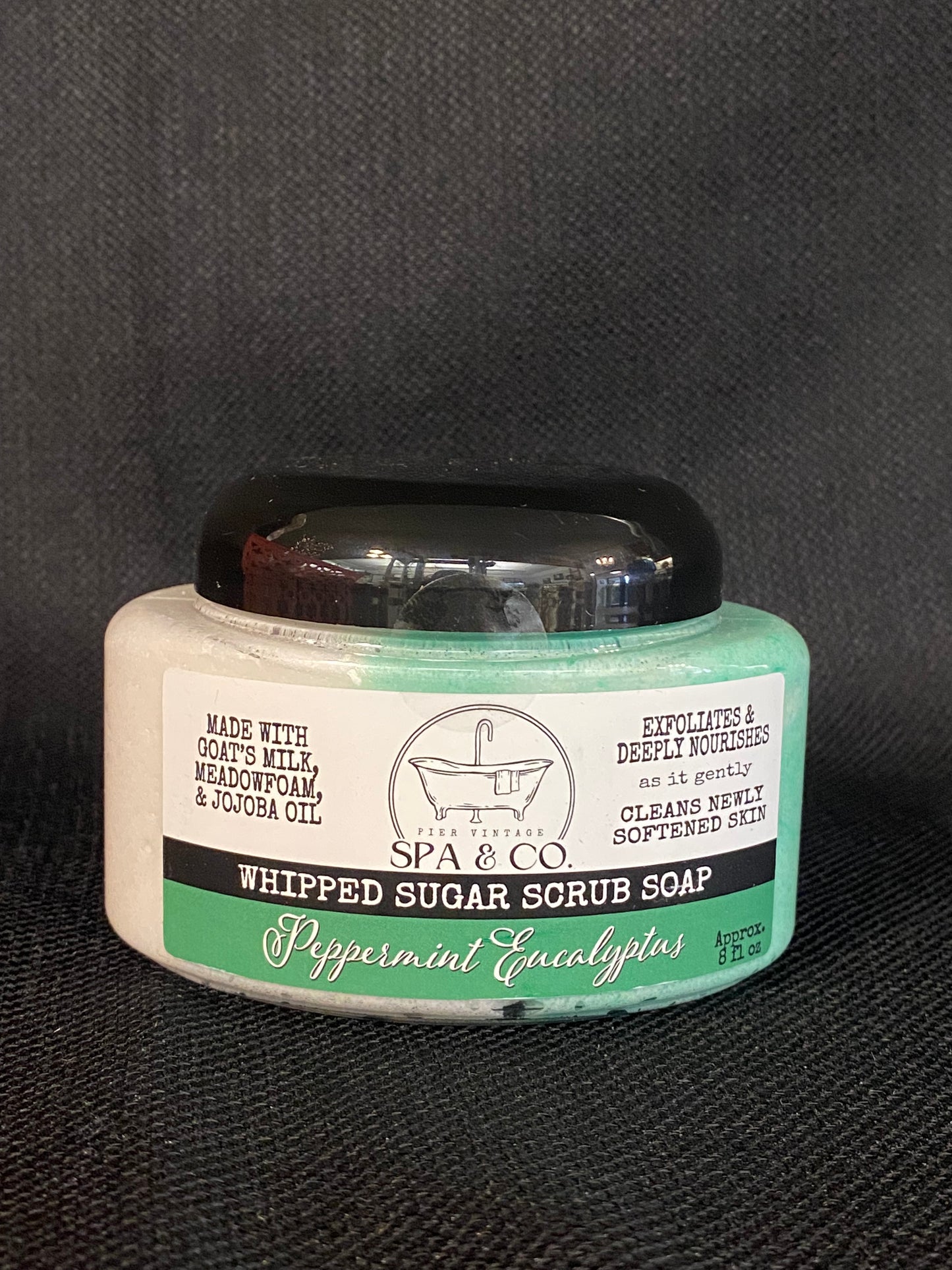 Jar of whipped sugar scrub soap with a green and white label on a textured black surface from Pier Vintage Spa & Co.