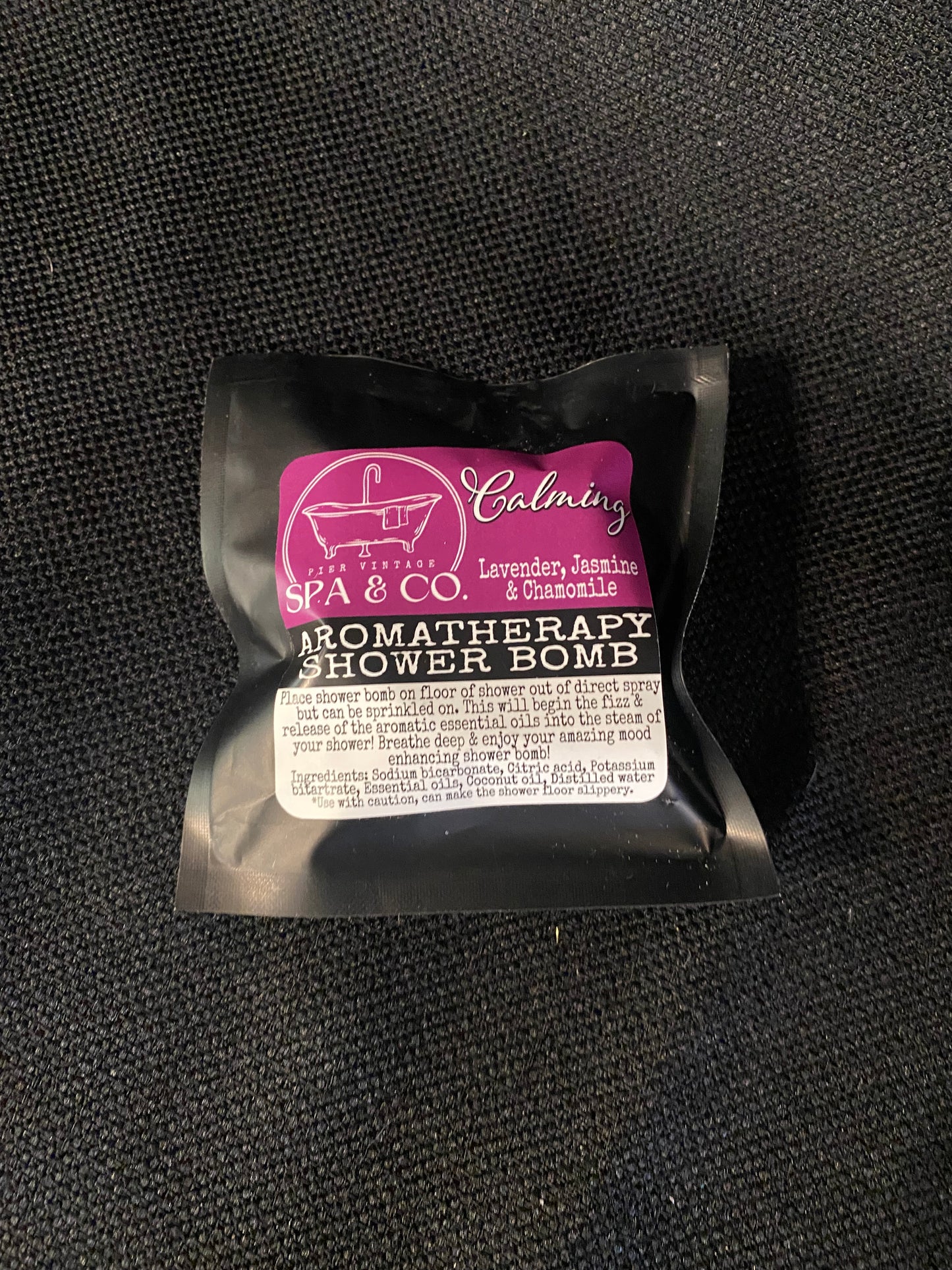 A small packaging of a shower bomb with visible branding on a black textured background