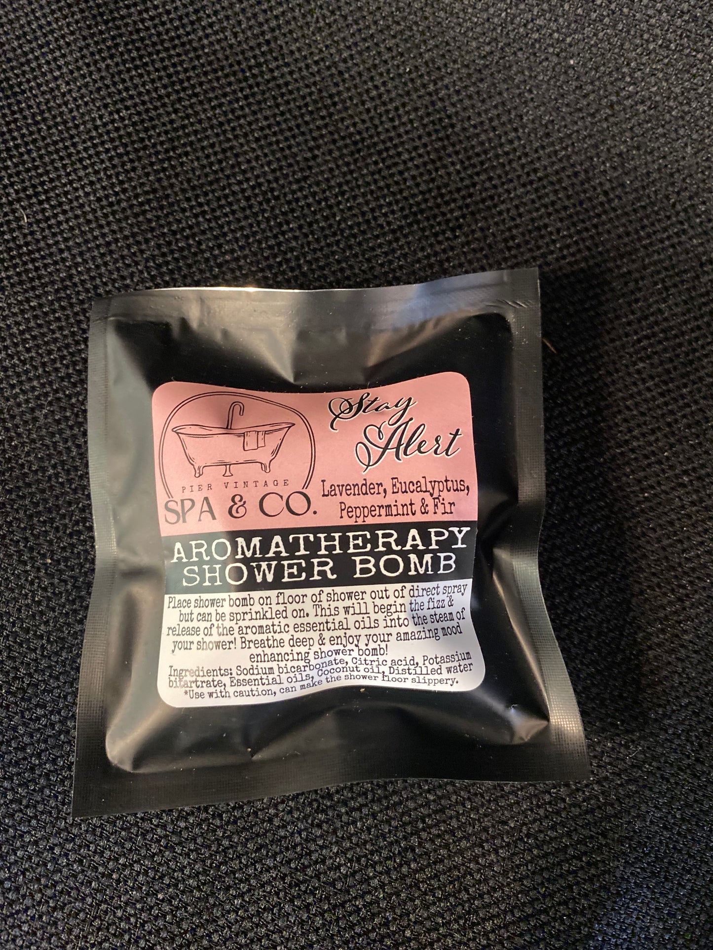 Aromatherapy shower bomb packaging with a pink label on a textured black background