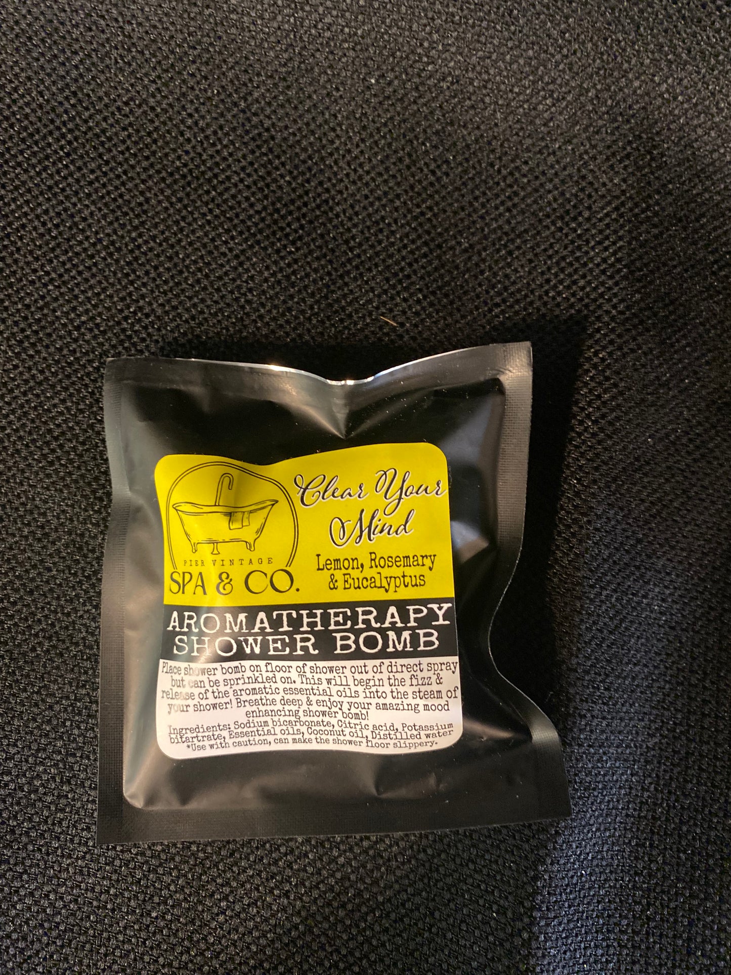 Aromatherapy shower bomb packaging with a yellow label on a black textured background