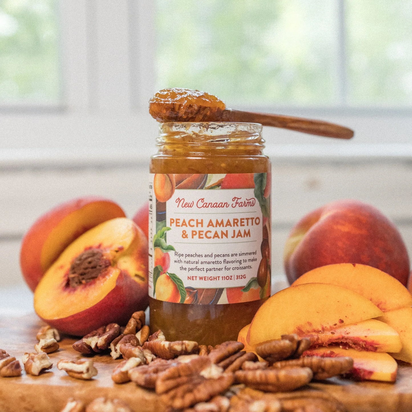 Jar of Peach Amaretto & Pecan Jam with a spoon, surrounded by peaches and pecans on a wooden surface.