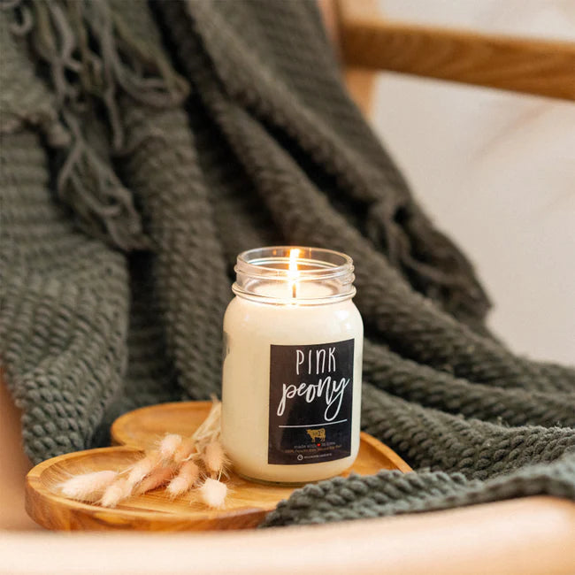 Candle in a jar labeled 'Pink Peony' on a wooden coaster with a green blanket in the background