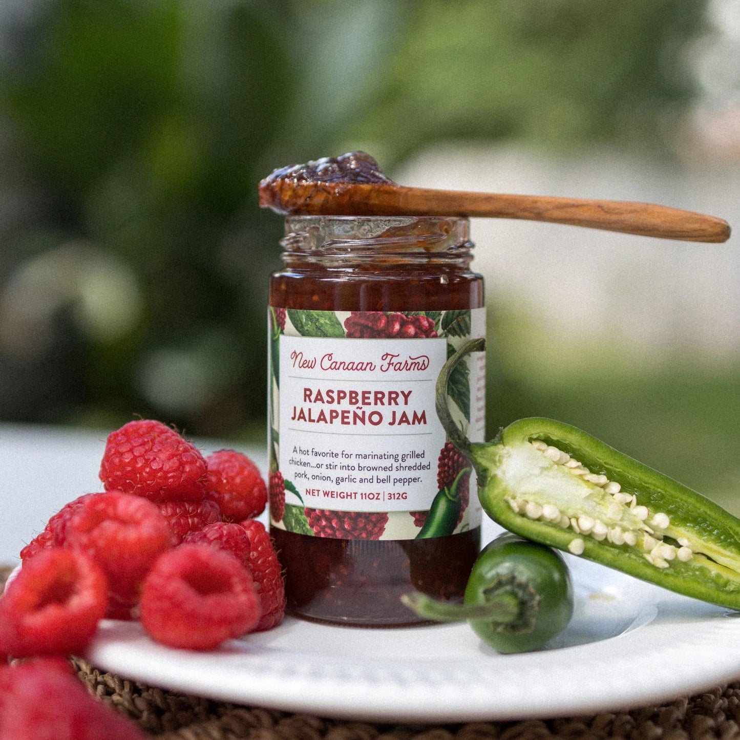 Jar of raspberry jalapeno jam with a wooden spoon, raspberries, and jalapenos on a plate.