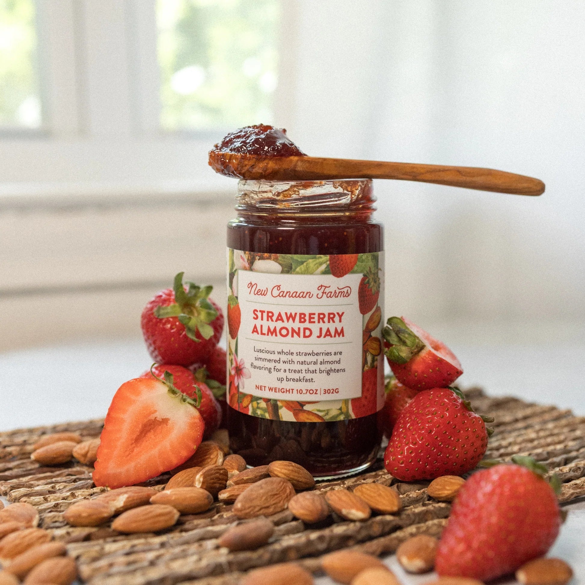 Jar of strawberry almond jam with a wooden spoon, surrounded by strawberries and almonds on a woven mat.