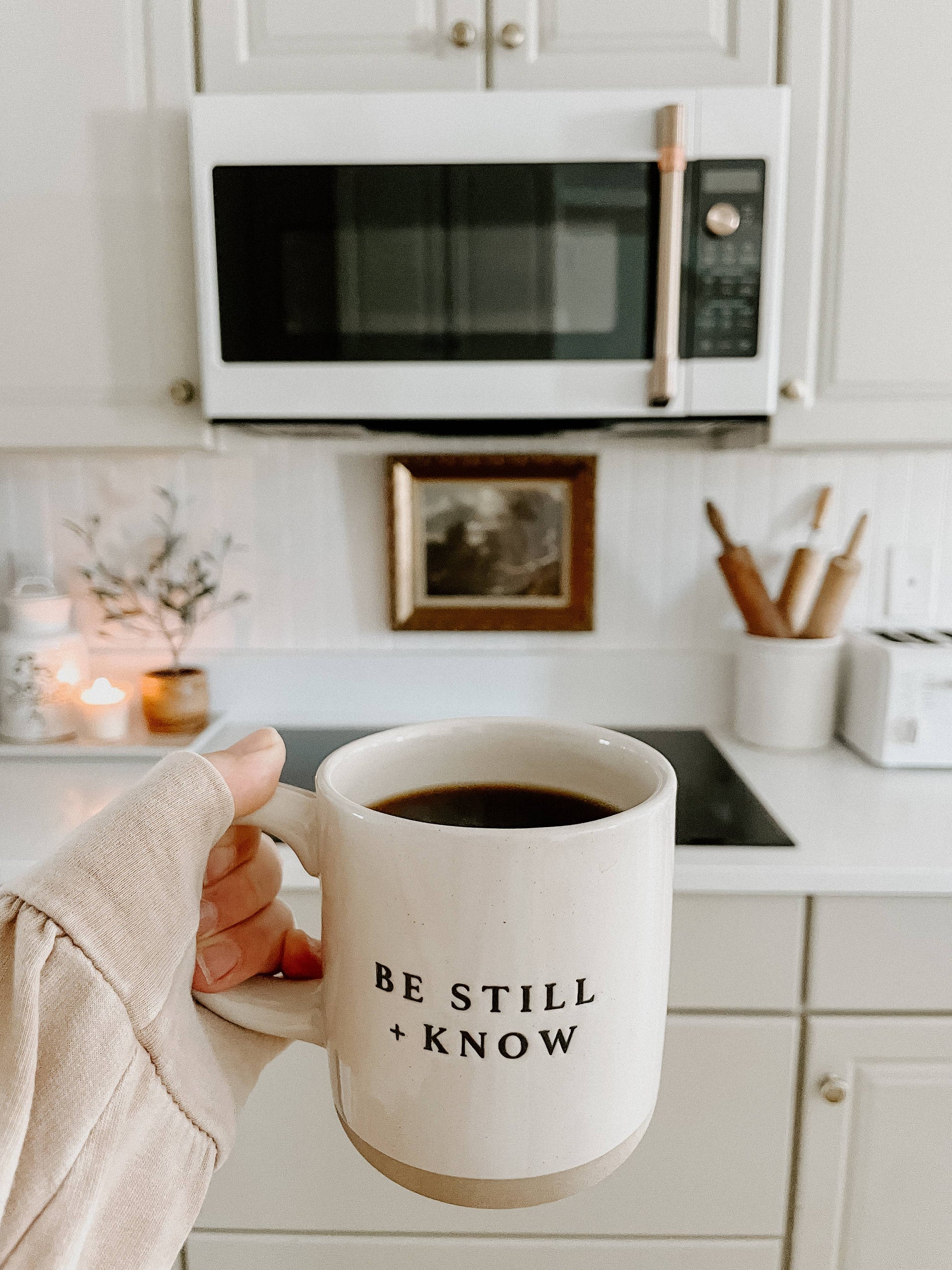 Person holding a mug with 'Be Still + Know' text in a kitchen.