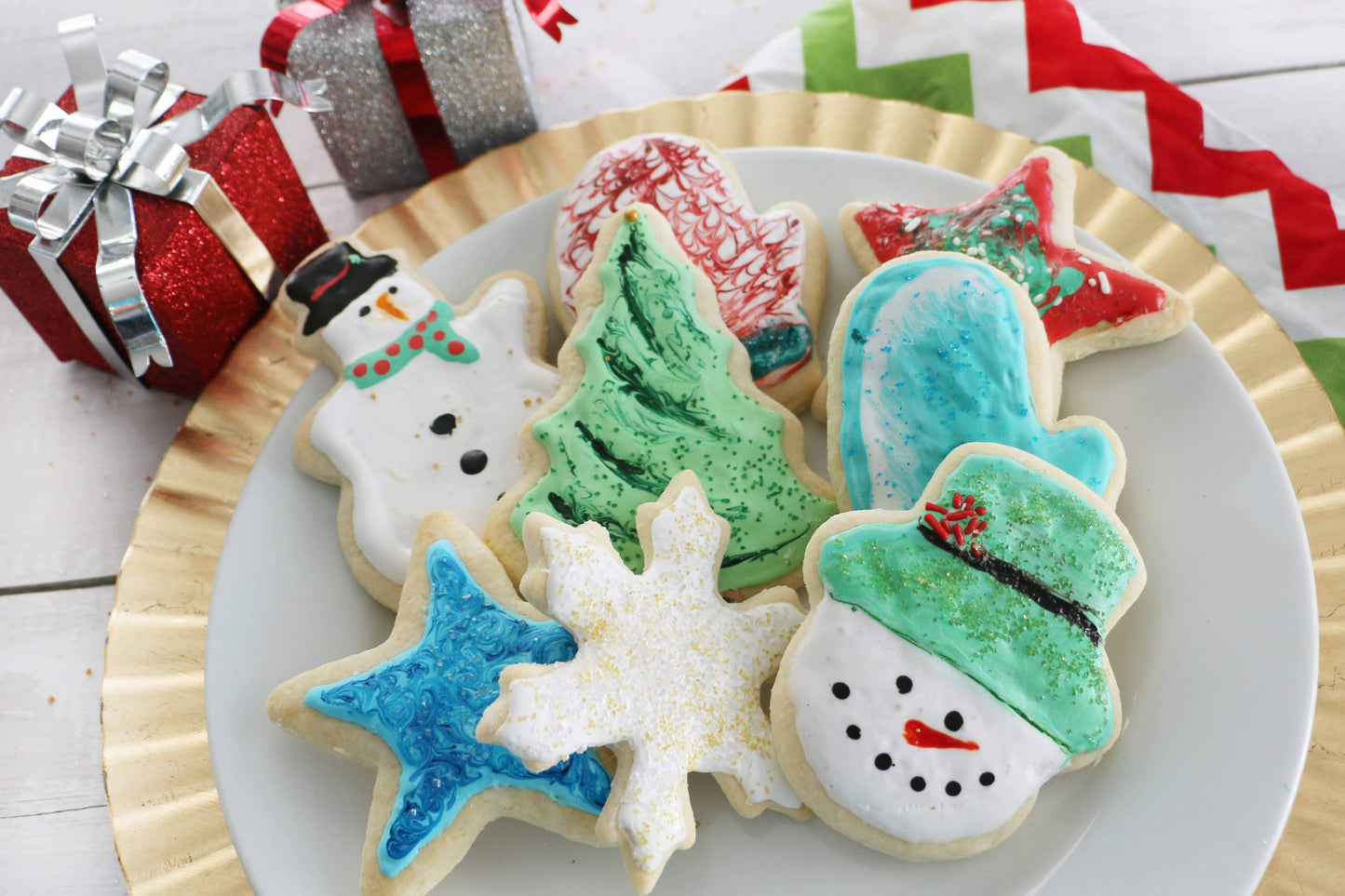 Decorative cookies shaped like a snowman, Christmas tree, and star on a plate with Christmas presents in the background.