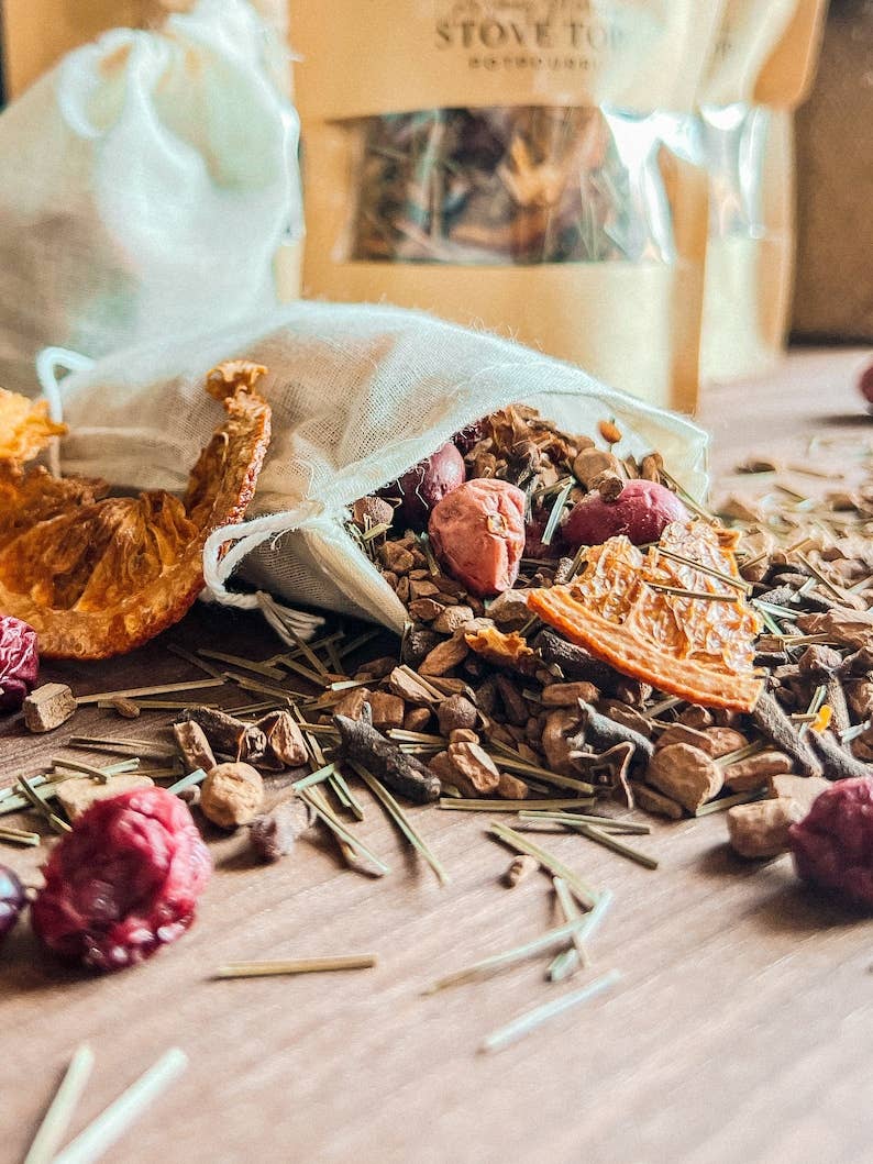 A close-up of loose-leaf herbal tea spilled from a small white mesh bag onto a wooden surface, with dried fruit, flowers, and green stems visible. A larger branded bag stands blurred in the background.