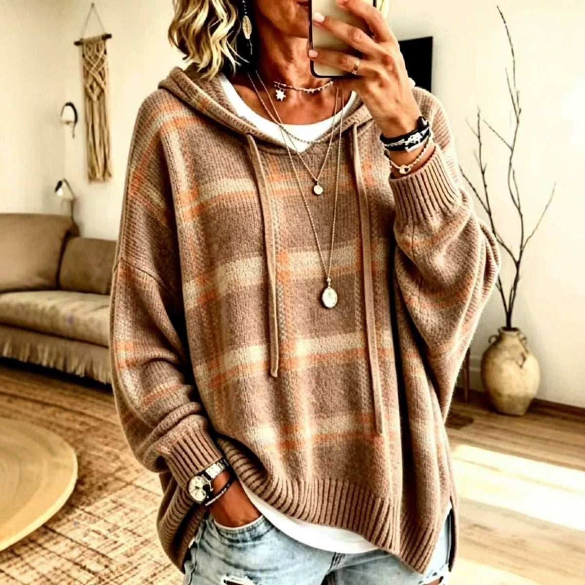 A woman takes a mirror selfie wearing an oversized brown and beige plaid knit hoodie with a white undershirt and light-wash jeans in a cozy living room setting.