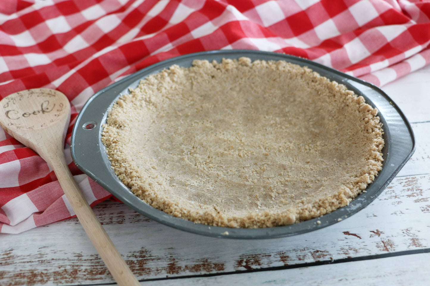Pie crust in a pan on a wooden surface with a red and white checkered cloth in the background.