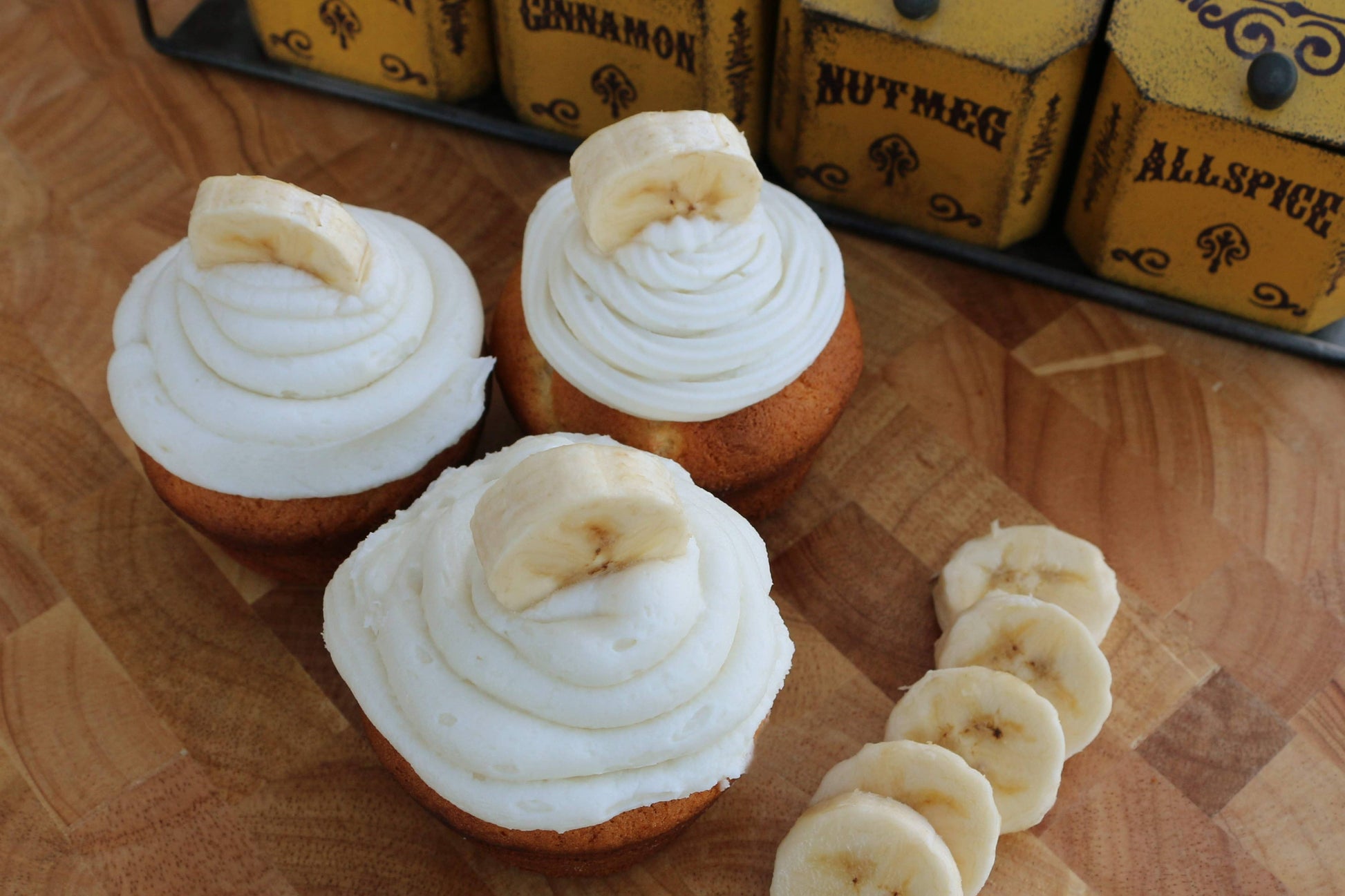 Three cupcakes with white frosting and banana slices on a wooden surface with spice jars in the background.
